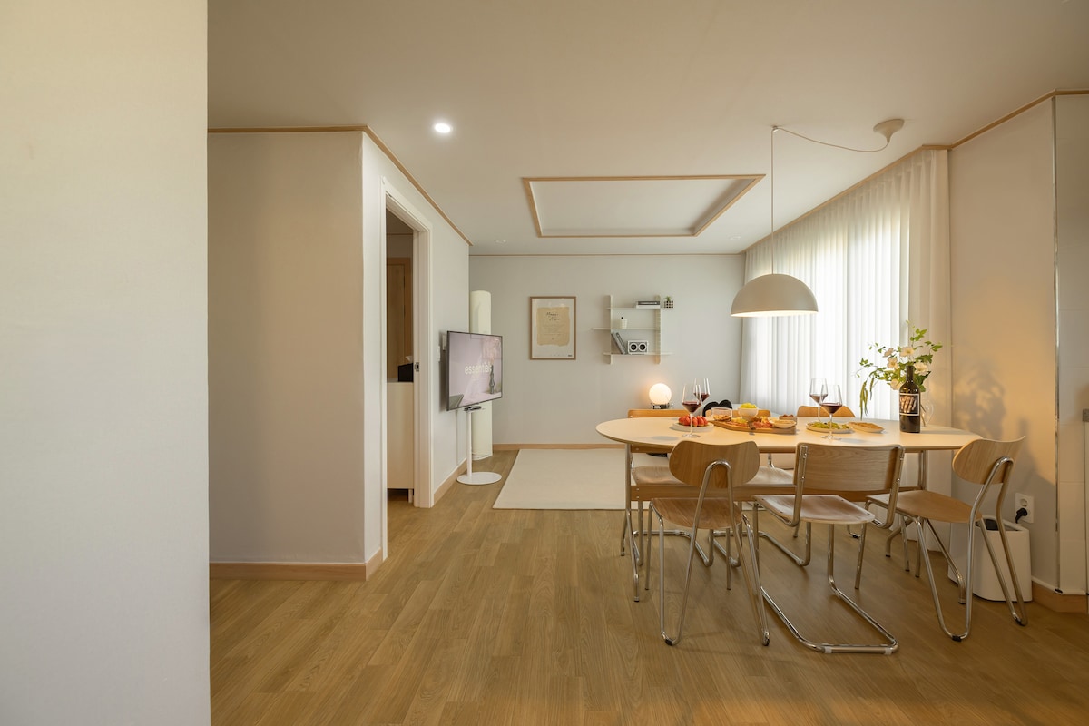 A bright dining area is shown, featuring a round table surrounded by wooden chairs. A stylish pendant light hangs above the table, which is set with an assortment of fruits. Soft natural light filters through sheer curtains, illuminating the space.