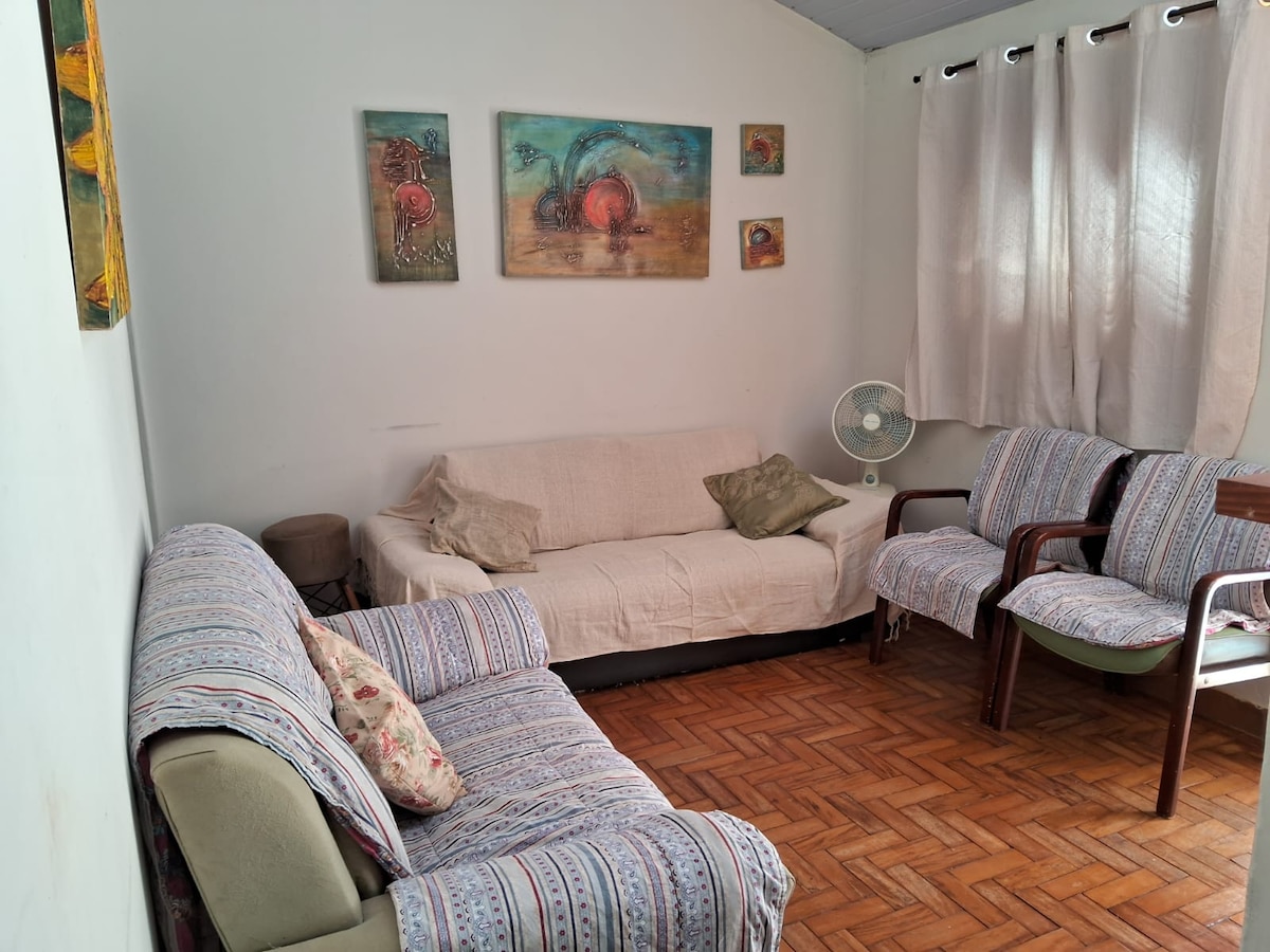 A living area displays a light-colored sofa with decorative pillows and two wooden chairs. The space is adorned with colorful wall art, and natural light filters through sheer curtains, creating a pleasant environment. A portable fan is positioned in the corner.