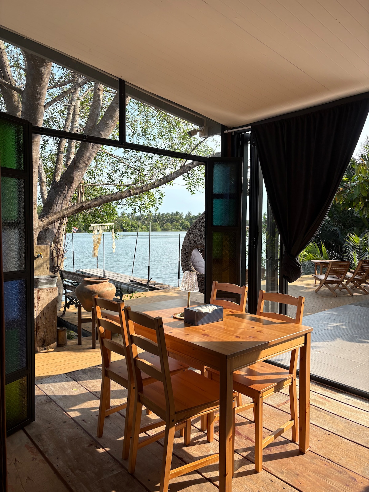 A dining area is shown with a wooden table surrounded by four chairs. Large glass doors open to reveal a view of the water and greenery. A tree is visible just outside, adding to the natural ambiance, while light streams through the colorful glass panes.