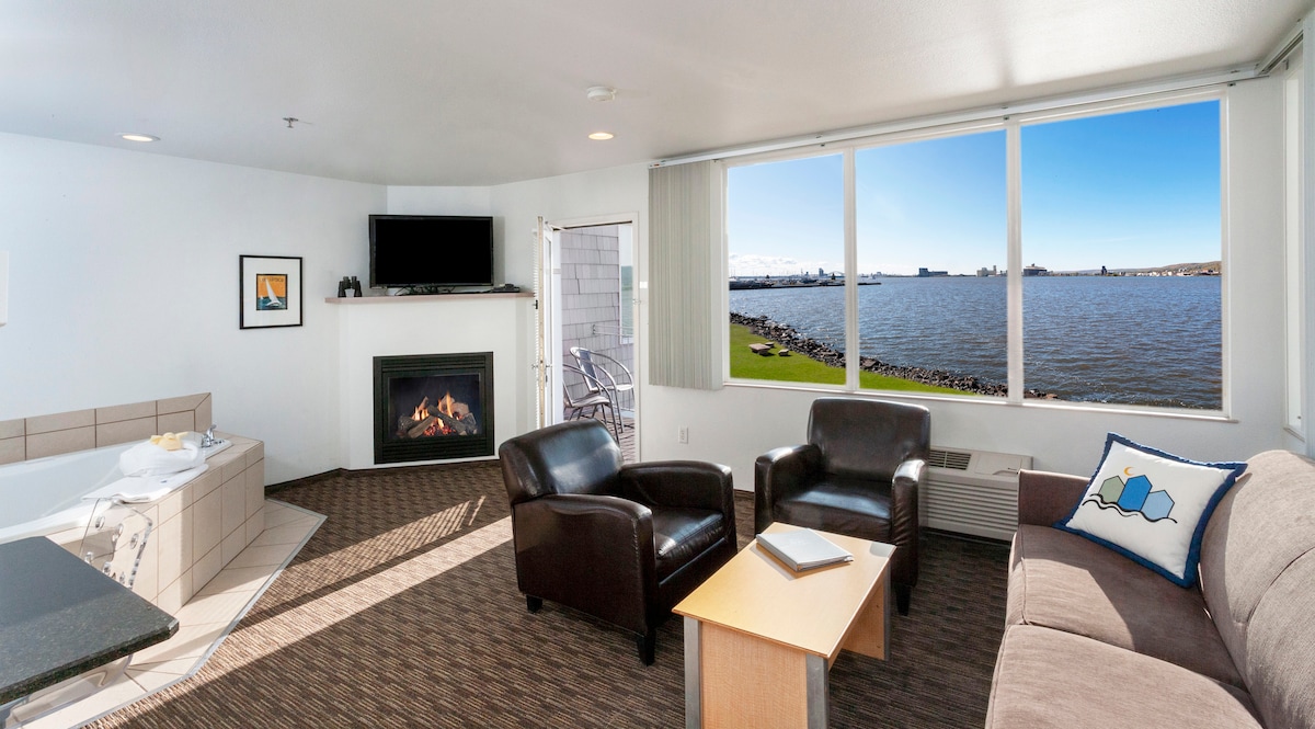 The living room features a cozy seating arrangement with two armchairs and a sofa, complemented by a small wooden coffee table. A gas fireplace is visible alongside a two-person whirlpool tub. Expansive windows provide a view of the water and outdoor space.