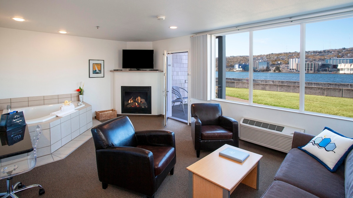 A spacious living area features a gas fireplace and large windows offering views of Duluth Harbor. Comfortable seating includes two black chairs and a beige sofa with decorative cushions. A whirlpool tub is positioned alongside a convenience center with a wet bar sink. A flat-screen TV is mounted on the wall.