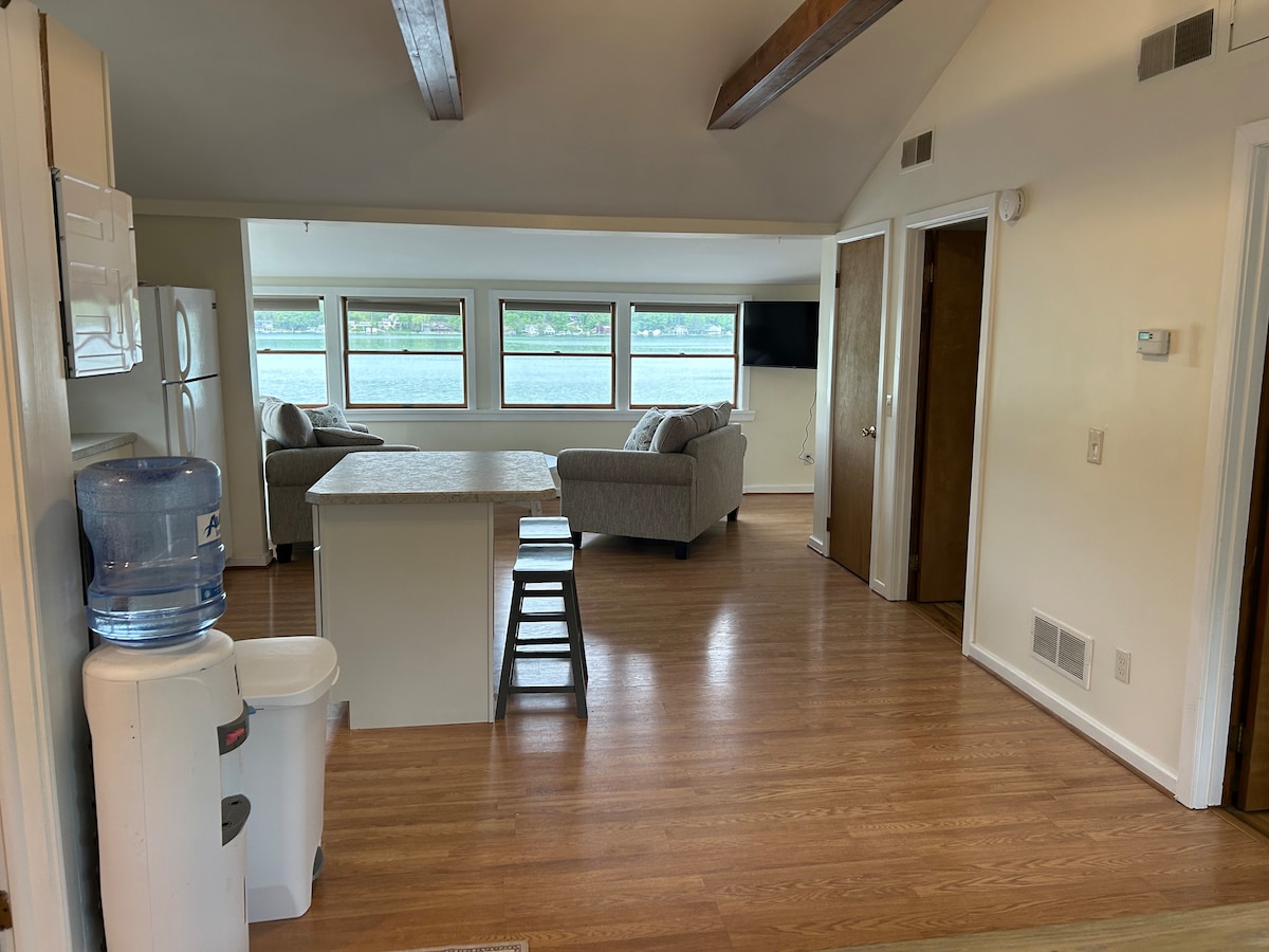 An open living space features natural light streaming through large windows with views of the lake. A modern kitchen area is equipped with a refrigerator and an island, while two comfortable seating areas are provided for relaxation. Wood beams accentuate the ceiling.