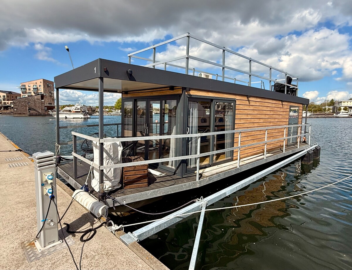 A modern houseboat is anchored at the marina, showcasing a wooden facade and large glass doors. A spacious rooftop terrace is visible, surrounded by railings, offering an inviting space to enjoy panoramic views of the water and surrounding area.