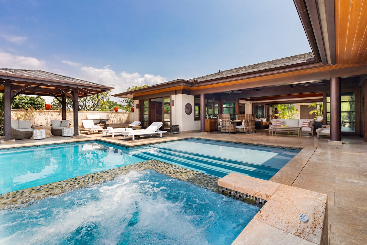 The outdoor space features a spacious heated pool and spa, surrounded by lounge chairs and elegant seating. A covered pergola offers shade, while large glass doors connect the interior living area to the pool area, emphasizing open-concept living.