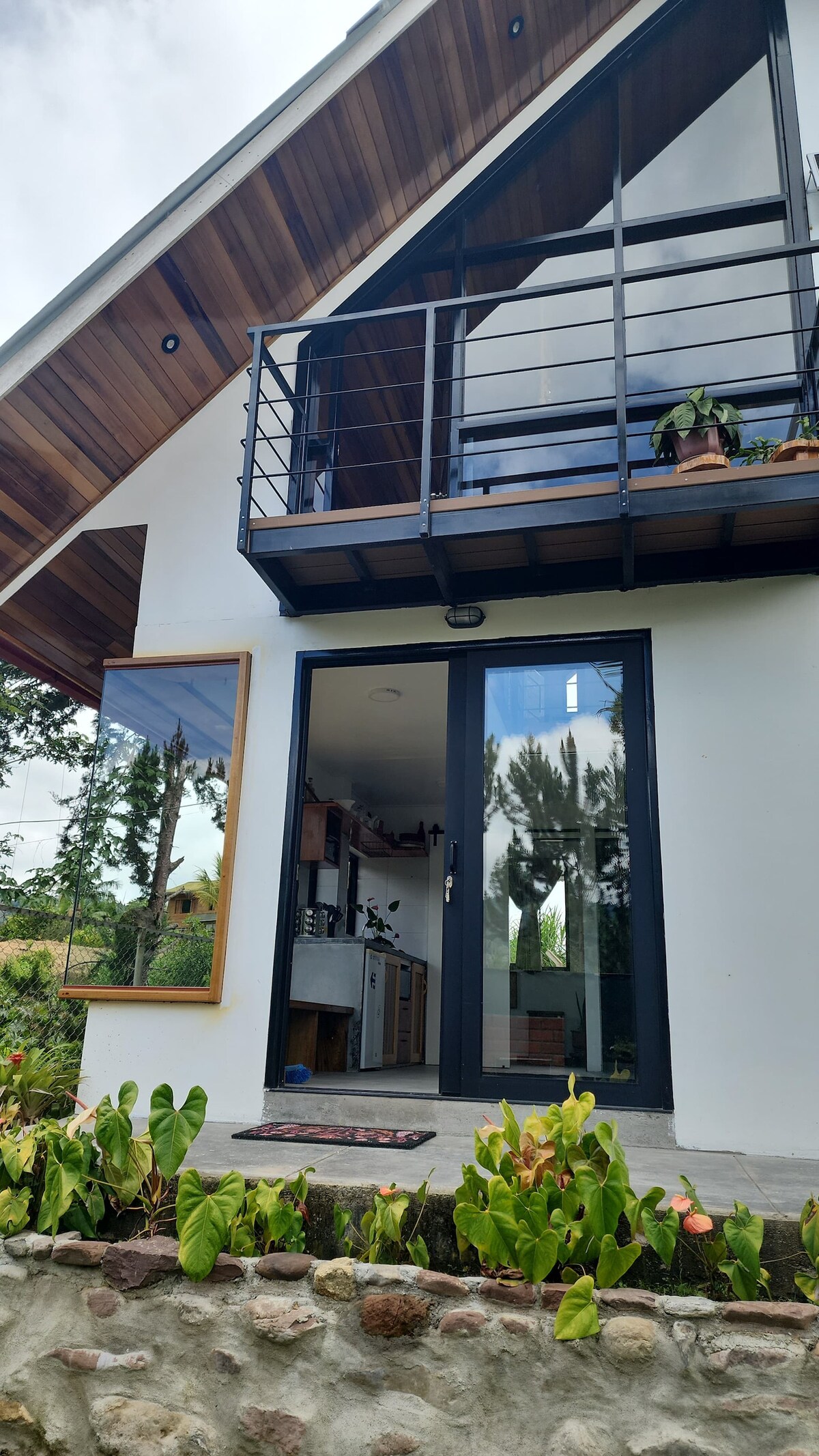The exterior features a modern design with a wooden overhang and black metal railing on the upper balcony. Large glass doors provide views into the interior space, while plants decorate the balcony and the entrance area is framed by greenery.