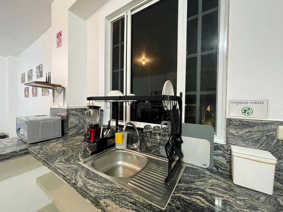 A kitchen area features a dark marble countertop with a stainless-steel sink. Kitchen appliances, including a blender and microwave, are visible. Natural light is provided by a large window, which offers a view of the night sky, while dishware is neatly organized on a rack.
