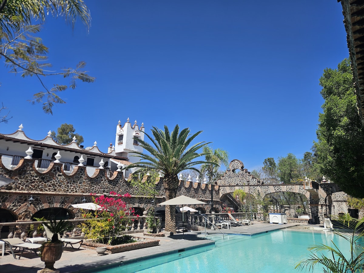 A spacious outdoor area features a clear swimming pool surrounded by stone structures and tropical plants. Palm trees provide shade near lounge chairs, while vibrant flowers add color to the stone architecture. A clear blue sky can be seen above, creating a serene atmosphere.