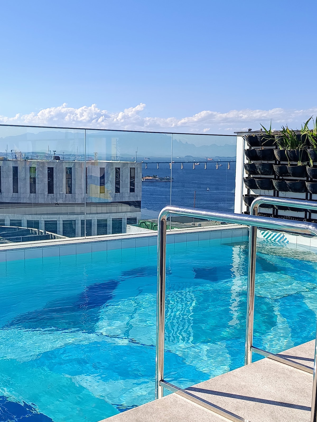 A rooftop pool is featured, surrounded by a smooth deck and glass railing. Expansive views of the water and distant mountains are visible, with a clear sky above. Greenery is present along the railing, contributing to the serene setting.