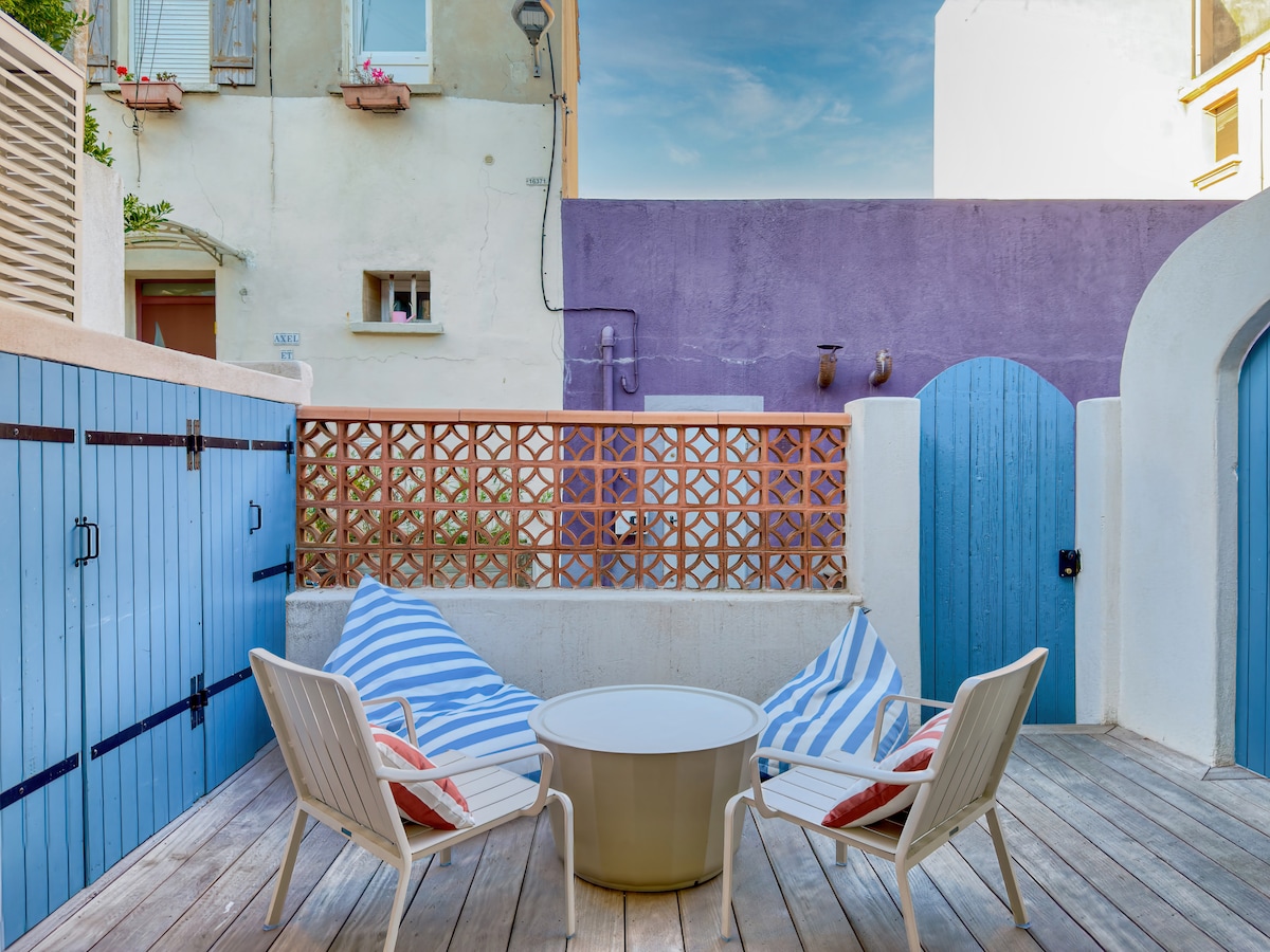 A private terrace features two lounge chairs with striped cushions and a round table at the center. The wooden decking creates a welcoming space, and colorful walls in the background add a charming ambiance.