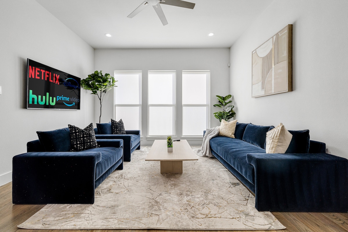 The living room is styled with a soft area rug and two navy blue sofas, offering a comfortable seating arrangement. A wooden coffee table is centered between the sofas, complementing the modern aesthetic. Natural light filters through three large windows, illuminating the space.
