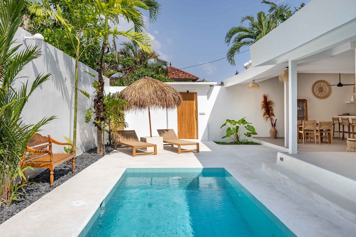 A serene outdoor space features a private pool surrounded by tropical plants. Two sunbeds are positioned next to the pool, while a thatched umbrella offers shade. The white walls and flooring enhance the bright atmosphere, contributing to a peaceful retreat.