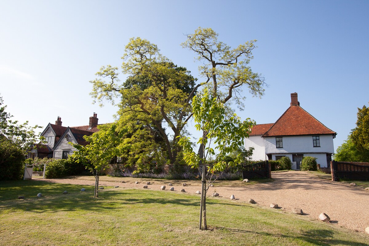 A spacious garden area is observed, featuring manicured lawns and a gravel driveway that leads to the cottage. Towering trees provide shade, while the architecture showcases distinct characteristics of traditional English cottage design, with a red-tiled roof and white walls.