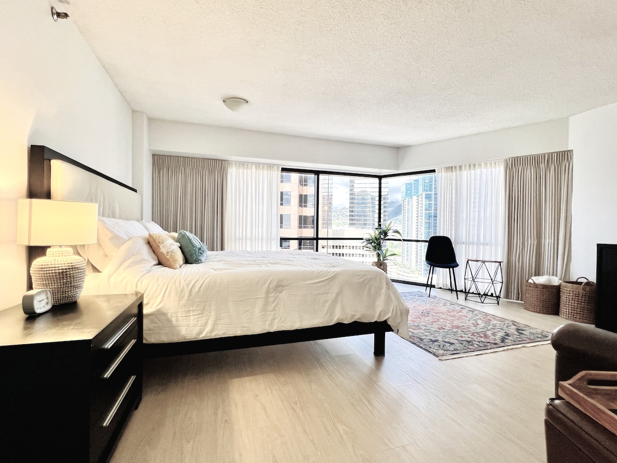 A spacious bedroom is furnished with a king-sized bed, complemented by light bedding and decorative pillows. Floor-to-ceiling windows allow natural light to illuminate the room and offer city views. A seating area is arranged thoughtfully, featuring a chair and a stylish table.