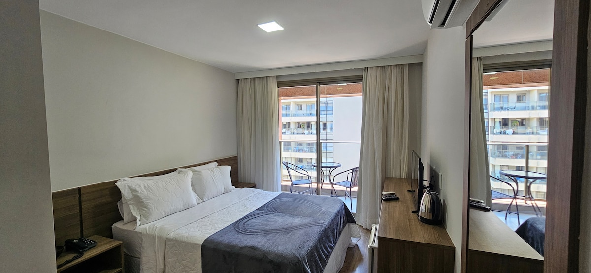 A bright and airy suite features a spacious bed with crisp white linens and a gray throw blanket. Large windows provide natural light and a view of the balcony, which is furnished with two chairs and a small table. A wall-mounted TV and a coffee maker are also present.