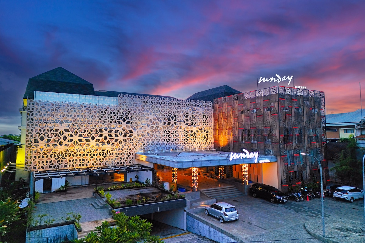The exterior of the Sunday Hotel Arshika is showcased against a vibrant sunset sky. A modern facade featuring intricate patterns is illuminated, with a welcoming entrance and a well-maintained landscape. Vehicles are parked in the driveway, emphasizing accessibility.
