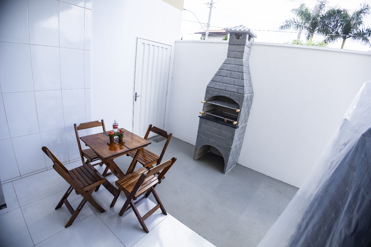 An outdoor area features a compact wooden dining table surrounded by four folding chairs. A stone barbecue grill stands against the white wall, providing a dedicated cooking space. The flooring is smooth with an open layout, allowing ample sunlight to enter.