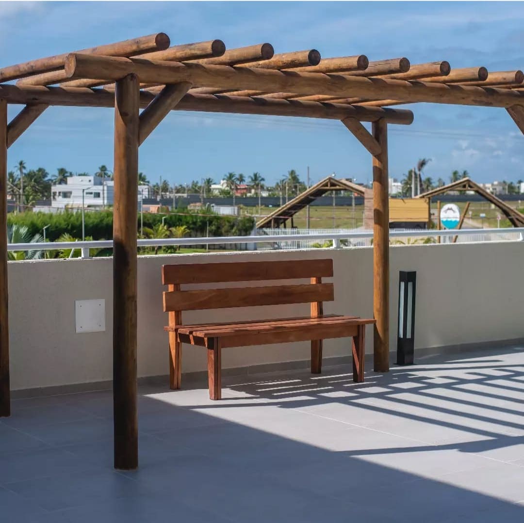 A wooden bench is positioned beneath a pergola, offering a shaded outdoor seating area. The tiled surface reflects sunlight from the clear sky, while greenery and structures are visible in the background, enhancing the outdoor experience.
