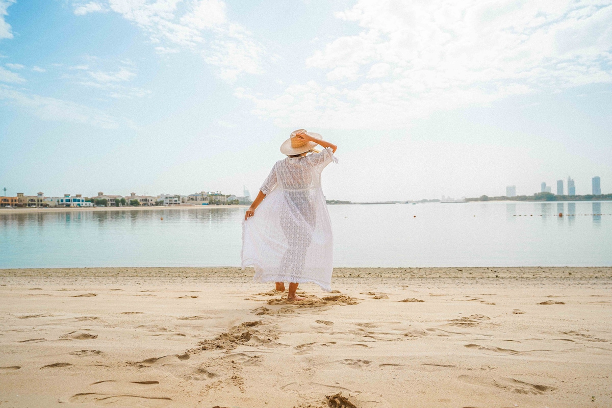 The image captures a serene beach scene with gentle waves lapping at the shore. A figure stands on the sandy beach, dressed in a light, flowing outfit and a wide-brimmed hat, facing the water. The tranquil surroundings and still water reflect the clear sky above.