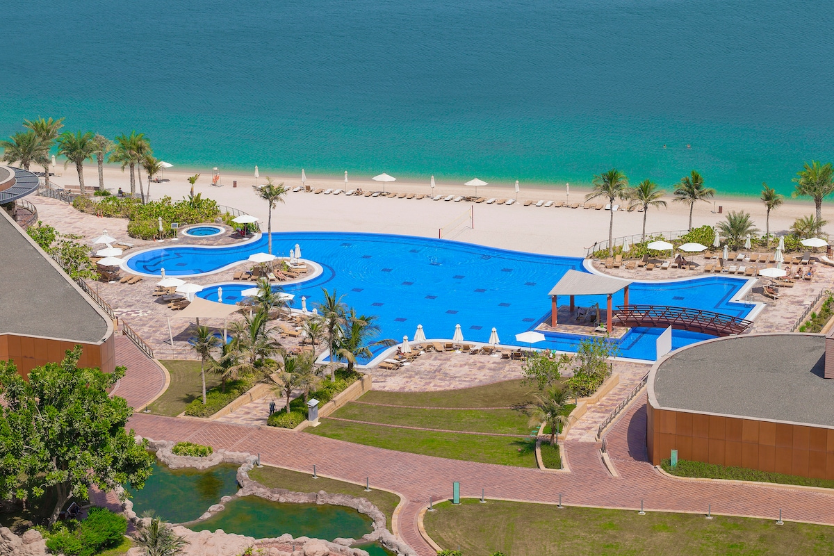 An expansive swimming pool is set against a backdrop of the beach and ocean, with sun loungers positioned along the shore. Lush palm trees surround the area, creating a relaxed environment. A shaded seating area is also visible near the pool.