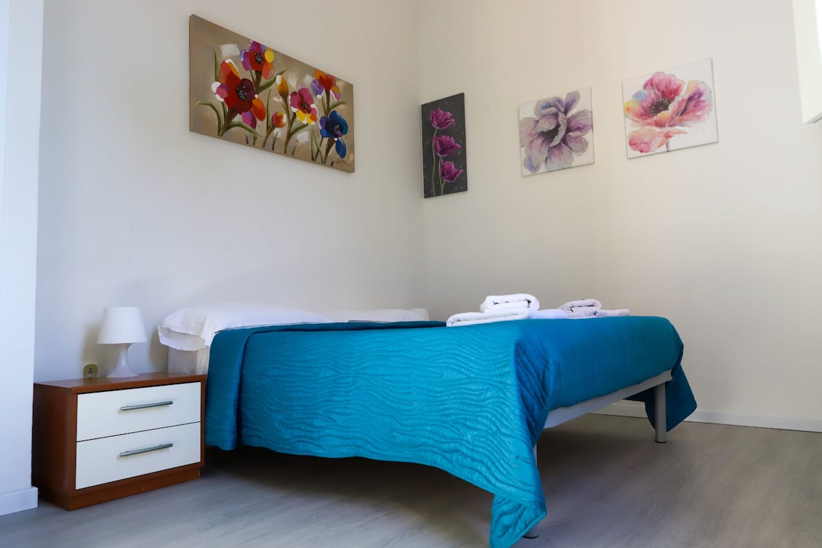 A bright bedroom features a spacious bed covered with a textured blue bedspread. Two neatly rolled towels are placed at the foot of the bed. The walls are adorned with floral artwork, adding a touch of color to the light, calming space.