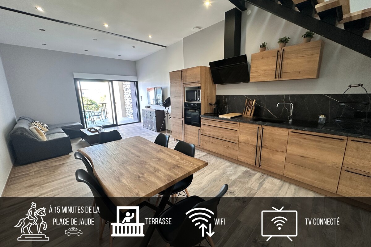 A modern kitchen and dining area features a wooden table surrounded by chairs, complemented by sleek cabinetry. A comfortable sofa is positioned nearby. Large glass doors allow natural light to fill the space, providing access to a terrace with outdoor seating.