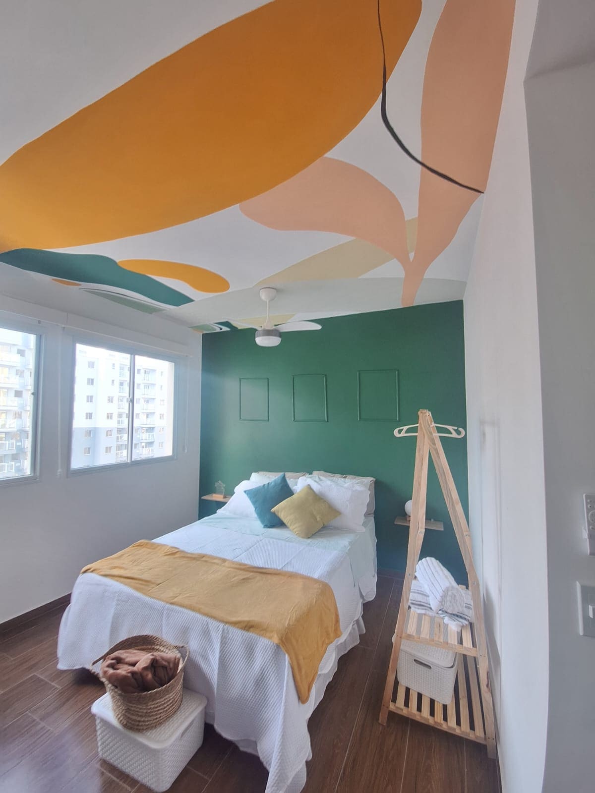 A bright bedroom features a green accent wall and a colorful ceiling design with organic shapes. A spacious bed is adorned with assorted pillows and a patterned throw. A simple clothing rack stands nearby, and large windows provide ample natural light.