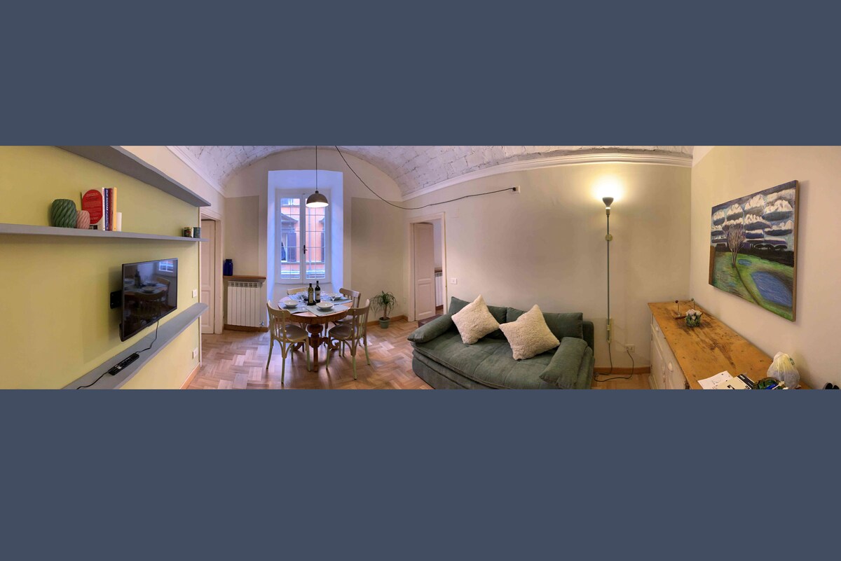 The living and dining area features a comfortable green sofa and a round dining table surrounded by chairs. A light fixture provides illumination in the space, with a TV mounted on the wall. An artwork hangs above a wooden sideboard, enhancing the room's ambiance.