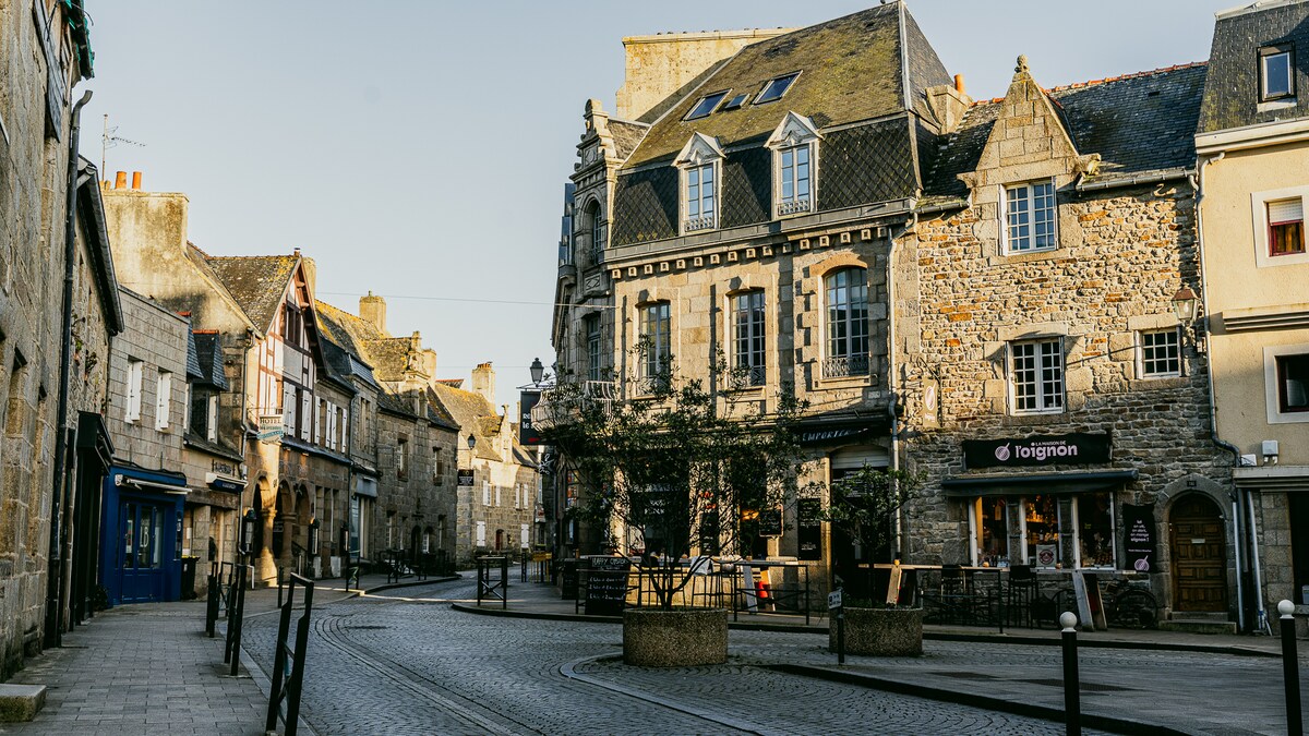 A charming street scene is depicted, featuring historic stone buildings with various architectural details. Shops and cafes line the cobblestone path, while trees and street lamps provide subtle greenery and lighting. The atmosphere suggests a peaceful day in a welcoming village.