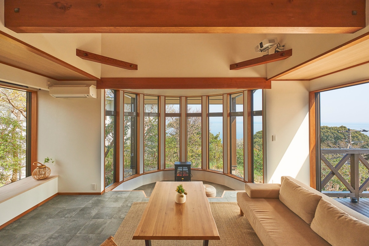 A bright and airy living space features large windows that offer views of the surrounding nature and distant ocean. A comfortable sofa is placed near a wooden table, and a projector is mounted on the ceiling. Natural light floods the room, enhancing its spacious feel.