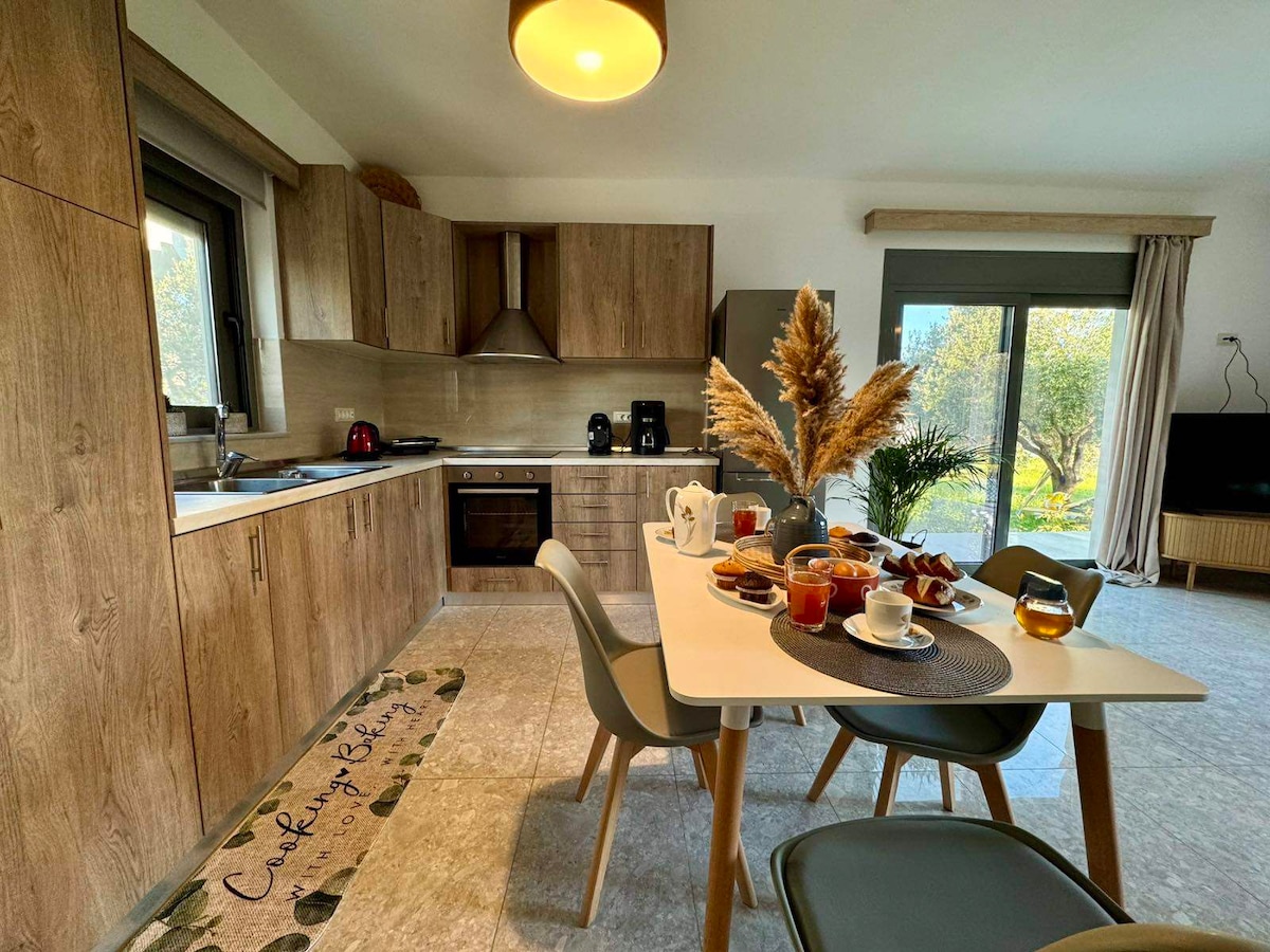 A modern kitchen and dining area features wooden cabinetry and a spacious table set for breakfast. A selection of pastries, beverages, and fruit is arranged invitingly. Large windows provide a view of the outdoor garden, enhancing the bright and airy atmosphere.