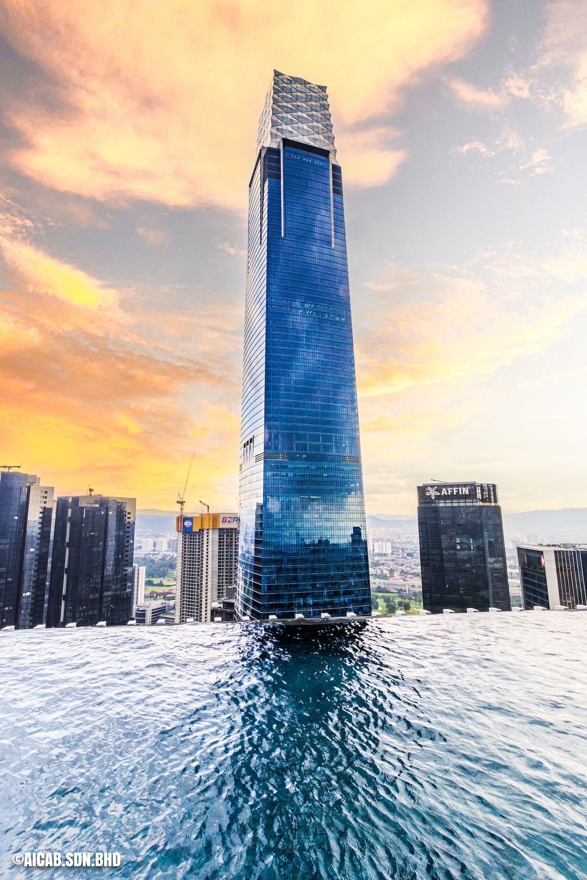 A tall skyscraper is featured prominently against a sunset sky, displaying a modern glass facade that reflects the vibrant colors. The scene captures the building’s height and surrounding cityscape, emphasizing its architectural design against the tranquil waters below.