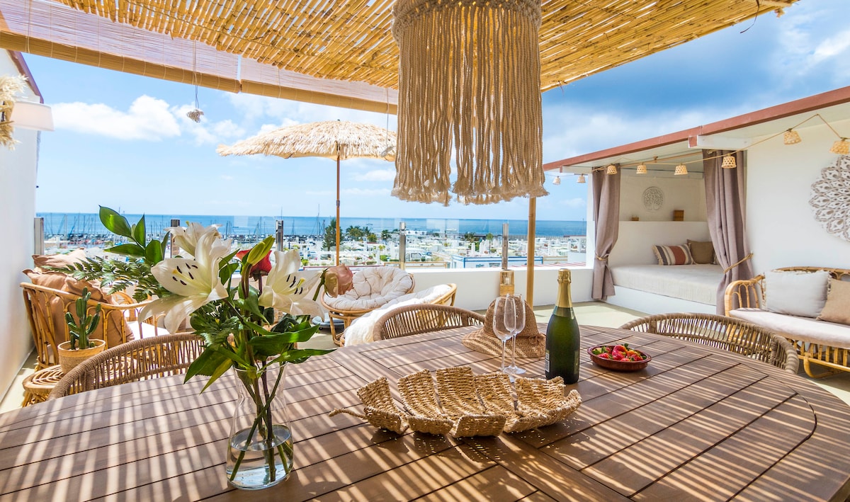 An outdoor terrace is adorned with natural wicker furniture, featuring a large dining table beside a cozy lounge area. Soft textiles and floral arrangements add a touch of warmth, while sweeping views of the sea are framed by a thatched roof overhead, creating a serene dining setting.