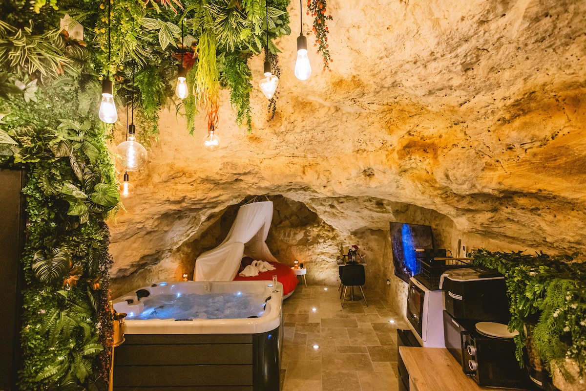 An intimate cave setting is adorned with foliage and soft lighting. A hot tub invites relaxation, while a bed draped in a white canopy is positioned against the rocky wall. A kitchenette with modern appliances is visible, enhancing the unique, tranquil atmosphere.