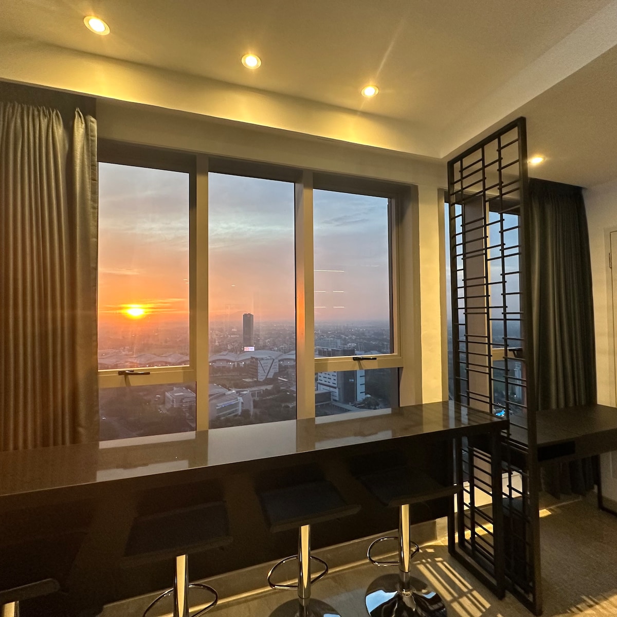 A spacious area is featured with large windows showcasing a glowing sunset view. A sleek bar table with high stools is positioned near the windows. Elegant drapes frame the windows, allowing natural light to enhance the warm ambiance.
