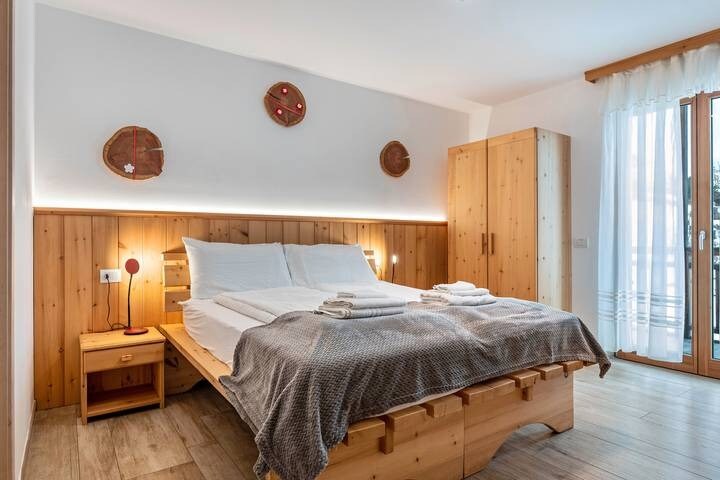 A spacious sleeping area features two neatly arranged beds with crisp white linens. Natural wood accents are present in the furniture, including a large wardrobe and a bedside table. Soft warm lighting is provided by wall-mounted lamps, with light filtering through sheer curtains.
