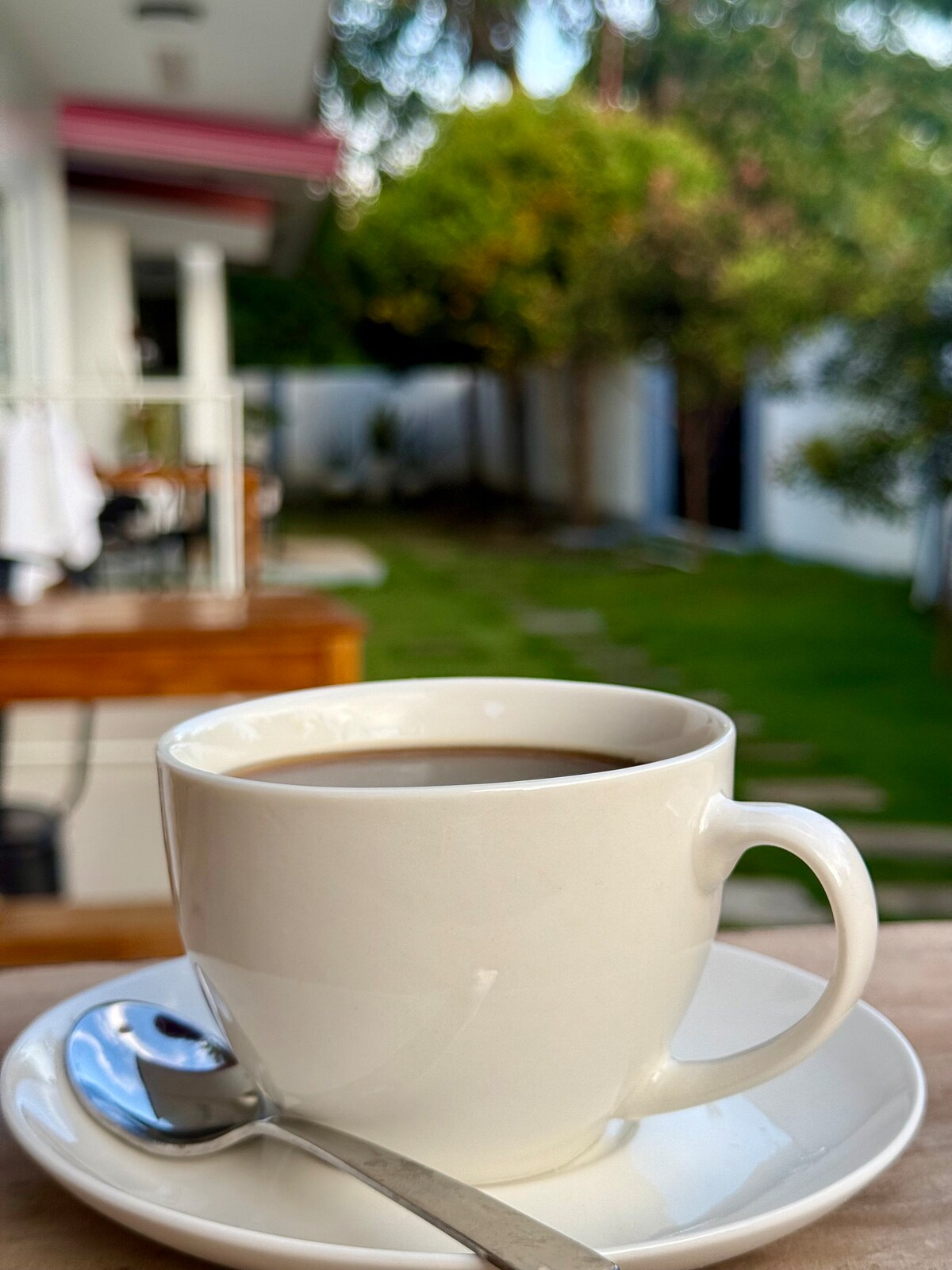 A white coffee cup is prominently displayed in the foreground on a saucer with a spoon beside it. The background reveals a lush garden surrounded by greenery, with a glimpse of a building and a patio area visible in soft focus.