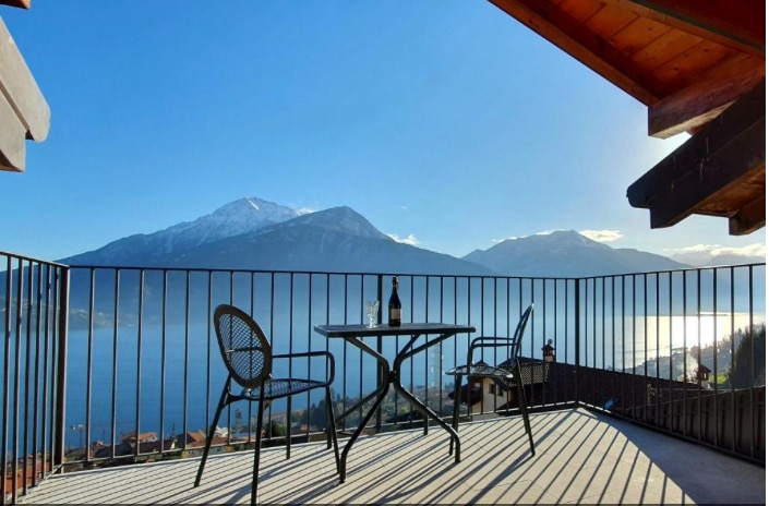 A balcony features a round table with two chairs, overlooking serene lake and mountain views. The space is sunlit, with the shadow of the railing creating a patterned effect on the floor. The natural scenery extends towards the horizon under a clear blue sky.