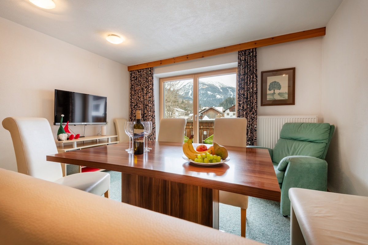 A dining area is presented with a wooden table surrounded by light-colored chairs. A bowl of fresh fruit sits at the center. A television is mounted on the wall, and large windows offer views of scenic mountains in the background, complemented by patterned curtains.