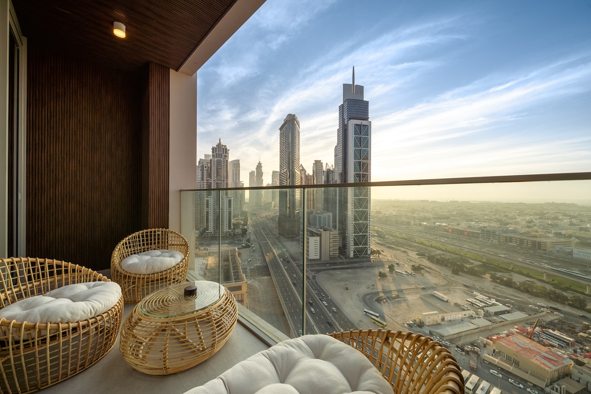A private balcony features modern woven seating with plush cushions, overlooking the expansive view of Downtown Dubai. The skyline, marked by high-rise buildings, is illuminated by natural light during sunset, creating a serene outdoor retreat.