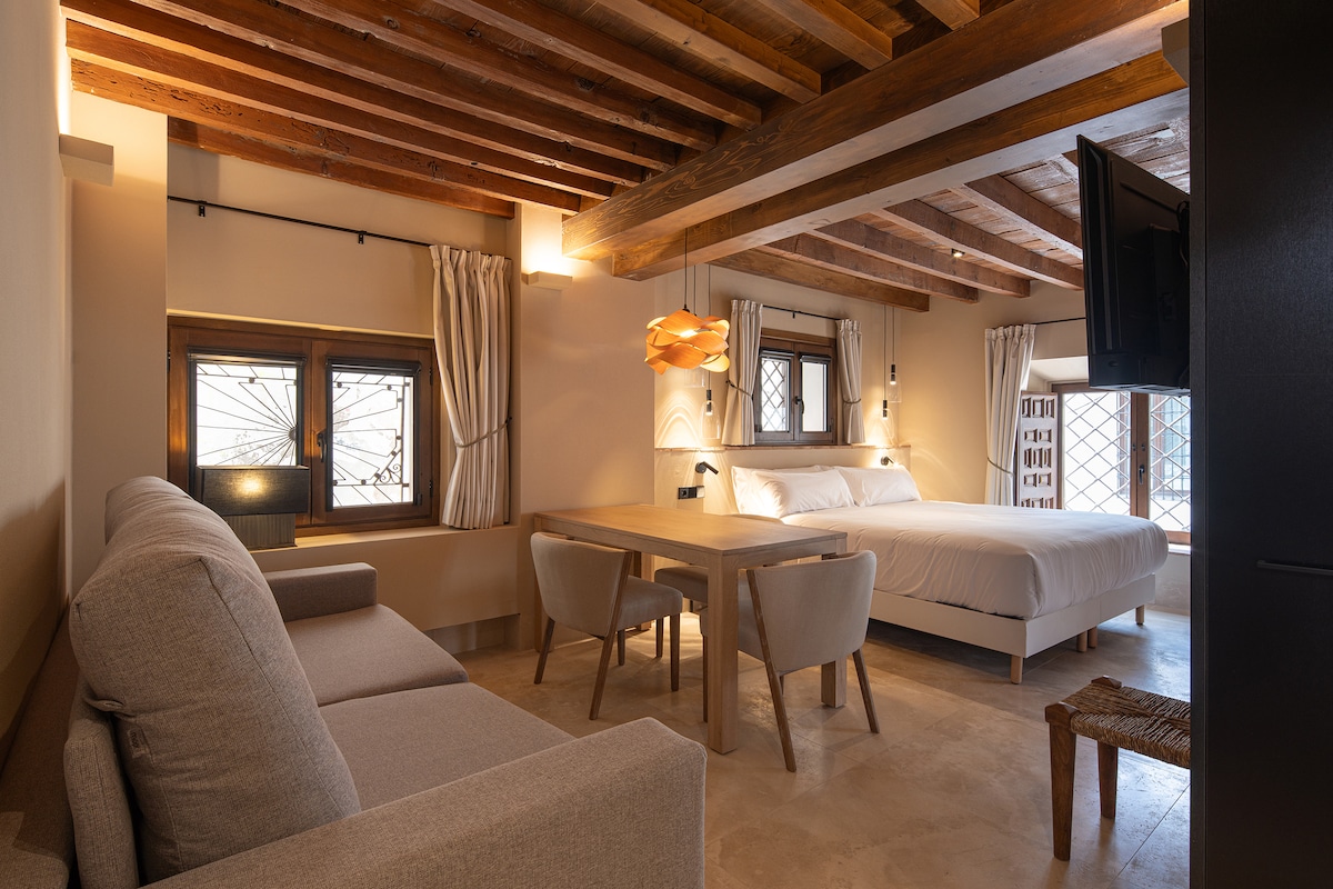 A spacious room features exposed wooden beams and soft lighting. A large bed is paired with a cozy sofa, while a dining table with light-colored chairs is positioned nearby. Natural light filters through two window lattices, enhancing the warm ambiance of the space.