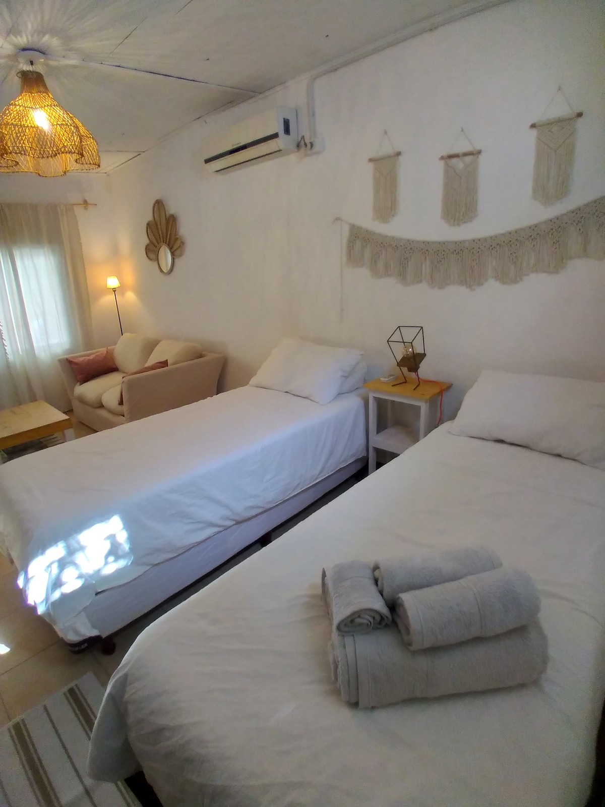 A well-lit sleeping area features two individual beds dressed in crisp white linens. A cozy seating area includes a beige armchair and a small side table. A decorative wall hanging and an ambient light fixture add a touch of character to the neutral-colored walls.