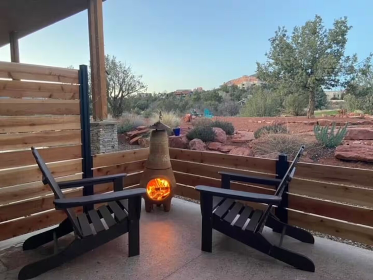 A private patio features two black Adirondack chairs positioned around a clay chiminea, which is glowing warmly. The backdrop includes natural red rock formations and desert vegetation, with a clear sky above, creating a tranquil outdoor space for relaxation.