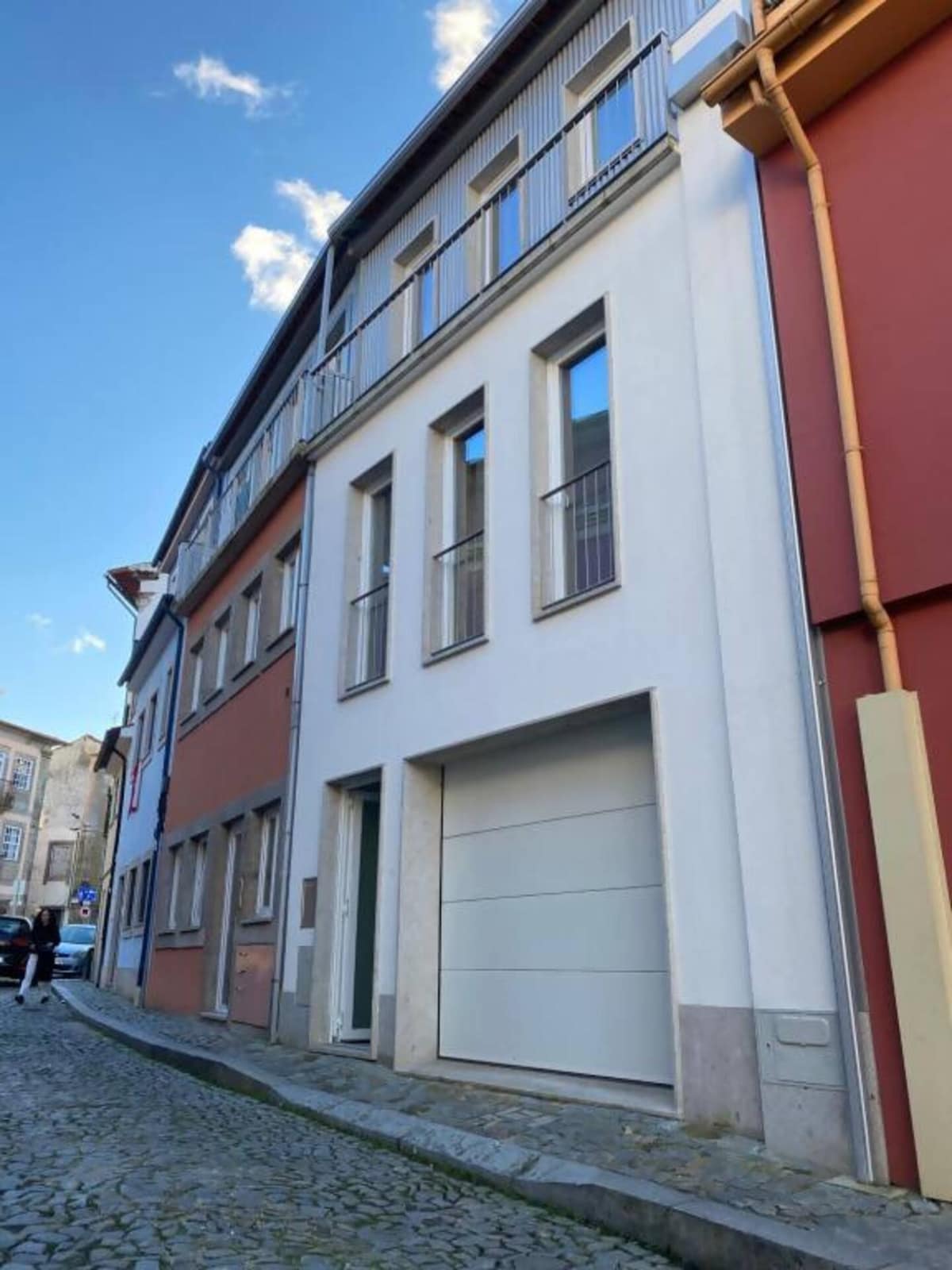 A modern building is displayed featuring a clean facade with multiple large windows. The structure is set along a cobblestone street, flanked by colorful adjacent buildings that add to the historic character of the area.