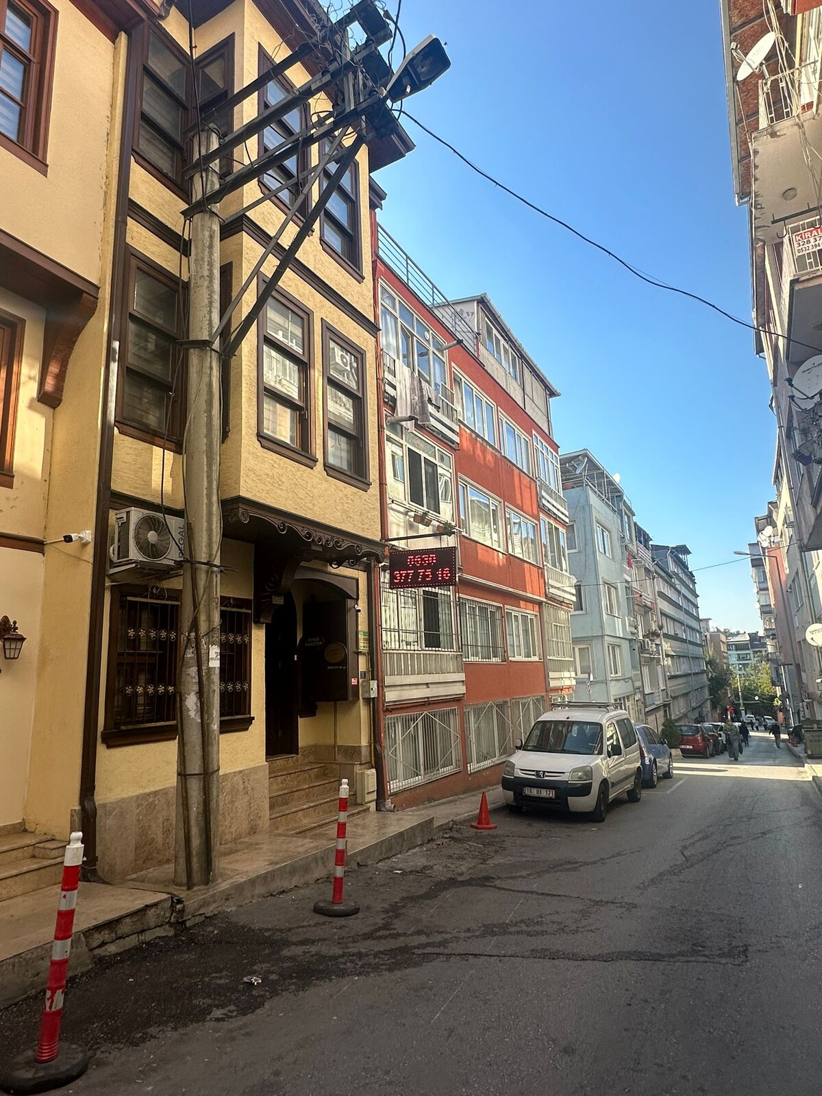A street view captures a mix of traditional and modern architecture, featuring a three-story building with large windows and a welcoming entrance. Nearby vehicles are parked along the side of the narrow road, with clear skies visible above.