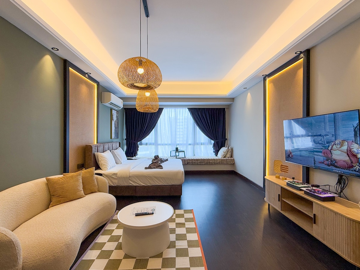 The studio features a spacious sleeping area with a premium king size bed, framed by elegant dark curtains. A comfortable seating area includes a stylish curved sofa and a circular coffee table. Natural light fills the room, highlighted by a large window with a plush bench.
