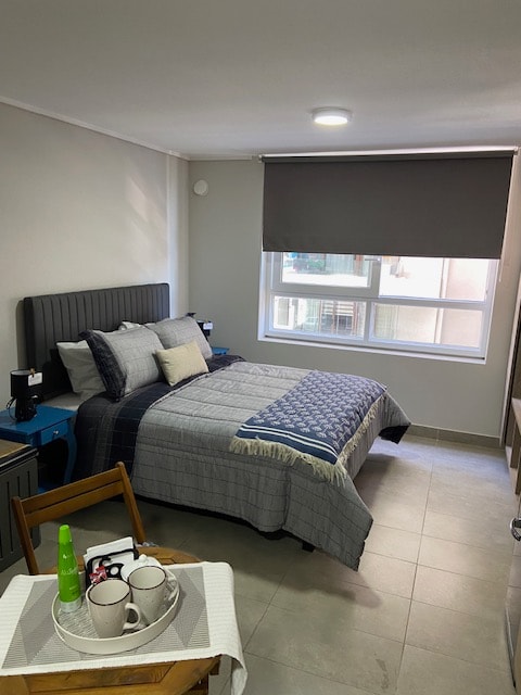 A well-organized studio space features a spacious bed with neutral-toned bedding and decorative pillows. A small table with two cups is positioned nearby, while a window with a gray roller shade allows natural light to filter in softly.