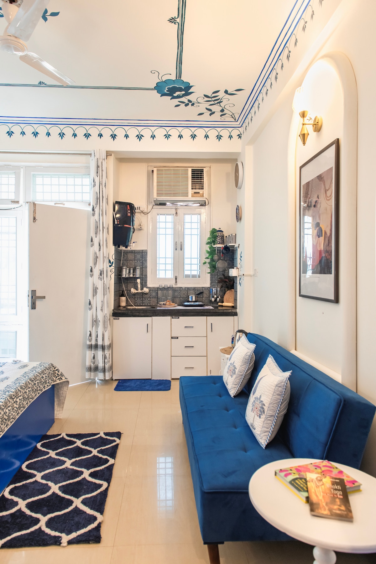 A cozy living area is presented with a blue sofa and decorative pillows, accompanied by a small side table. The kitchenette is visible, featuring dark countertops and white cabinetry. Decorative wall art and floral motifs add a charming touch to the space, with ample natural light illuminating the area.