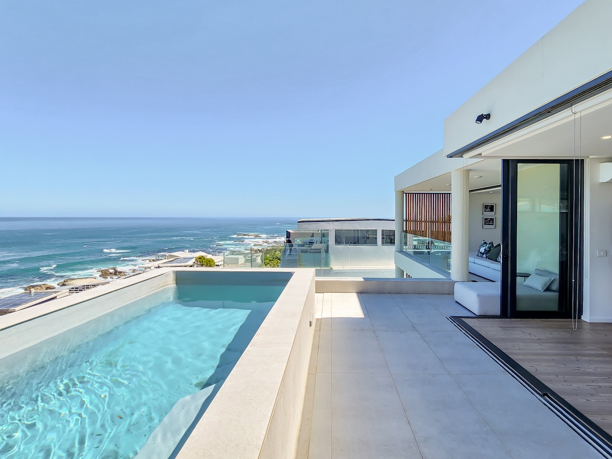 A sunlit terrace offers a view of the ocean, featuring a lap pool with clear water. The adjoining area is equipped with sun loungers, enhancing the outdoor space. Large glass doors provide access to the interior, allowing for unobstructed sea views.
