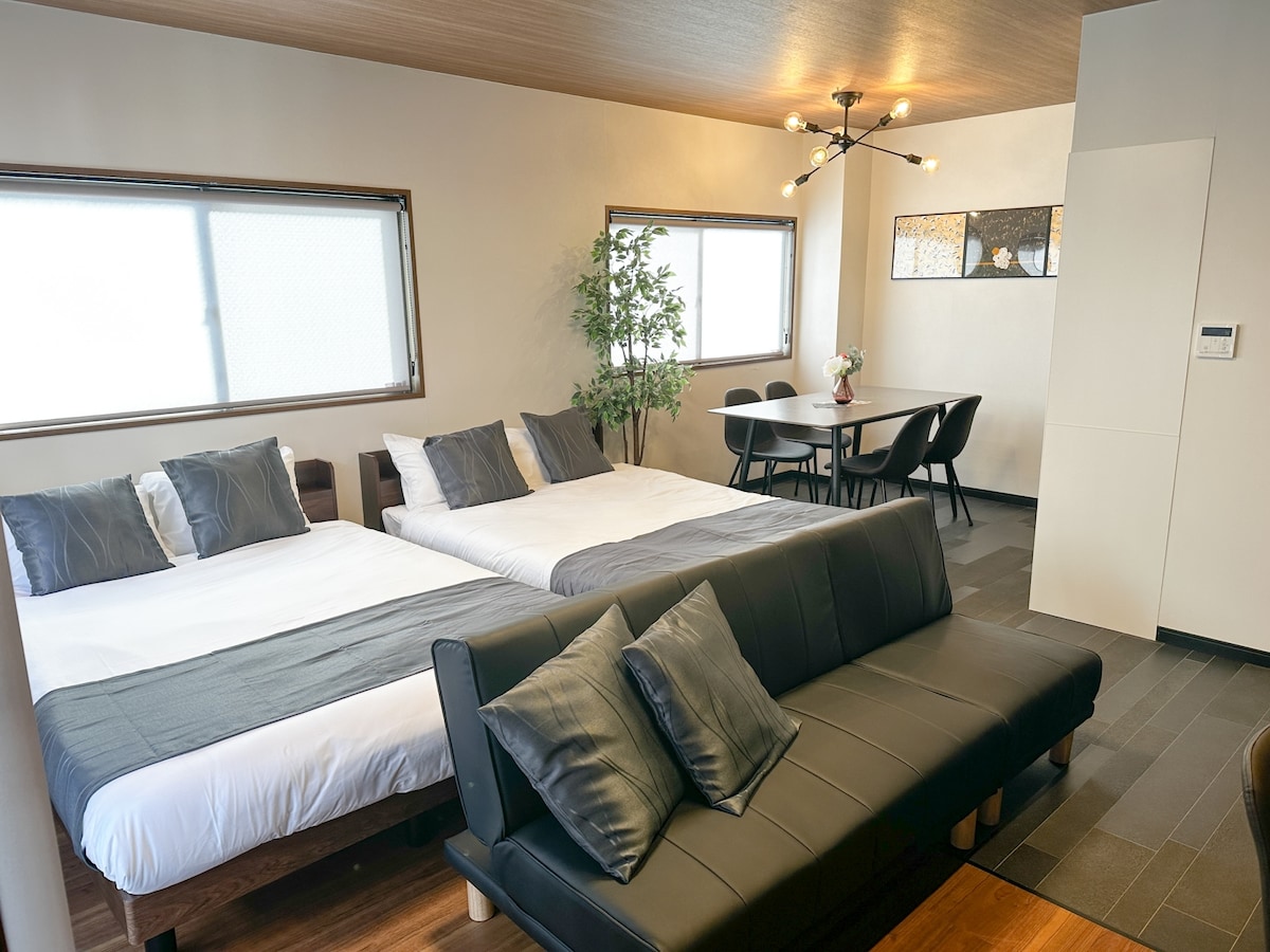 A spacious room features two large beds with dark gray bedding, complemented by a sizable sofa in front. A dining area with a round table and four chairs is visible, while large windows allow natural light to fill the space.