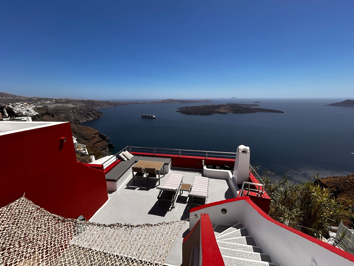 A spacious terrace overlooking the Caldera features outdoor seating areas with a dining table and lounge chairs. Steps lead down toward shared swimming facilities. The panoramic views capture the serene seascape and volcanic landscapes under a clear blue sky.